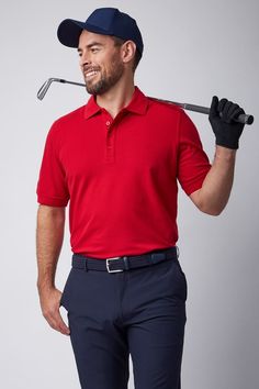 a man in a red shirt holding a golf club and wearing a blue hat while standing next to a white wall