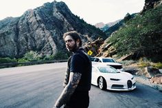 a man with tattoos standing in front of two cars on the road next to mountains