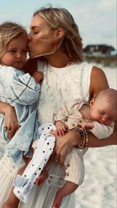 a woman holding two small children on her lap and kissing the baby's cheek