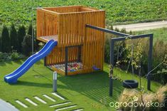 a wooden play structure with a blue slide in the grass next to some bushes and trees