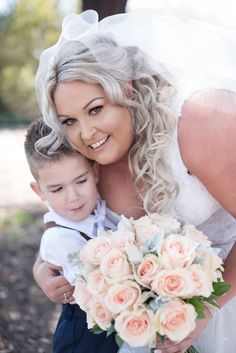 a woman in a wedding dress hugging a young boy with flowers on his lap and smiling at the camera