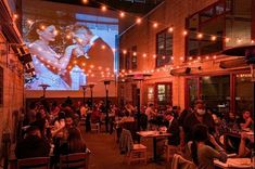 people sitting at tables in an outdoor dining area with lights strung from the ceiling and large screen on the wall