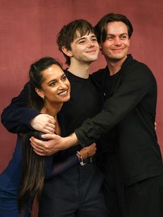 two men and a woman hugging each other in front of a red wall with their arms around one another