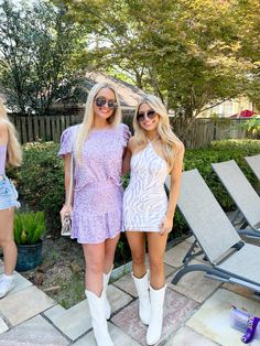 two women standing next to each other in front of some lawn chairs and trees with one woman wearing white cowboy boots