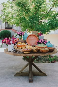 a wooden table topped with lots of food next to a lush green leafy tree