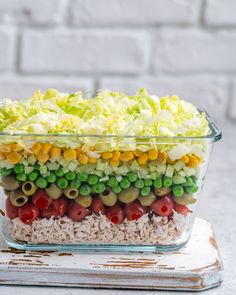 a layered salad in a glass dish on top of a wooden table with white brick wall