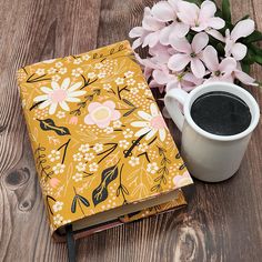 a cup of coffee next to an open notebook with flowers on it and a pen