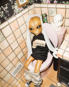 an alien doll sitting on top of a toilet in a bathroom next to a sink