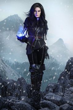 a woman dressed in black and holding a blue light up glove standing on top of a snow covered mountain