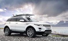 a white range rover with surfboards on top parked by the water in front of mountains