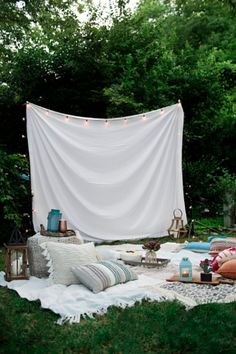 an outdoor seating area with pillows, blankets and lights on the grass in front of trees