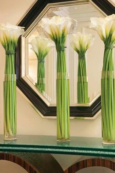 three vases with white flowers sitting on a glass table in front of a mirror