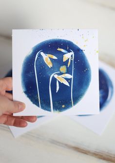 a hand holding up a card with three flowers on it and watercolor paint splatters