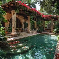 a pool surrounded by greenery and red flowers in front of a house with an outdoor gazebo