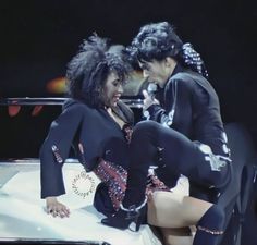 two women in black outfits sitting on top of a white car and one is holding a microphone