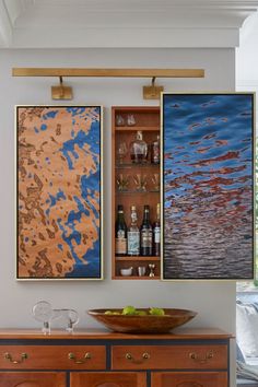 two paintings hanging on the wall above a wooden cabinet with wine glasses and fruit in it