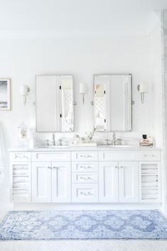 a white bathroom with double sinks and mirrors on the wall next to an area rug