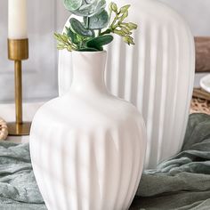 two white vases with plants in them sitting on a table next to a candle