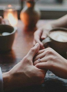 two people holding hands while sitting at a table with coffee cups and candles in the background