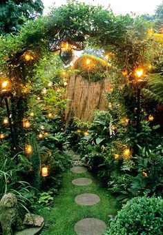 a garden with lots of plants and lights on the path leading to an archway that is surrounded by greenery