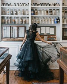 a woman in a black dress is standing near shelves with jars on them and looking at the floor