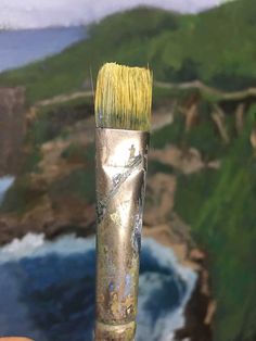 a brush with yellow bristles is held in front of an ocean and cliff background