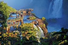 people are standing at the edge of a waterfall with a bridge that crosses over it