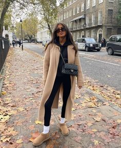 a woman is standing on the sidewalk with her hand in her pocket and wearing a tan coat