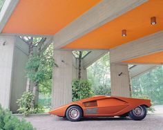 an orange car is parked under some concrete pillars in front of trees and bushes on either side of the building