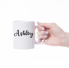 a person holding a white coffee mug with the word ashley on it in black ink