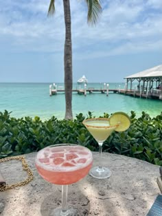 two cocktails sitting on top of a cement table next to the ocean and palm trees