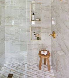 a bathroom with white marble walls and flooring, including a wooden stool next to the shower