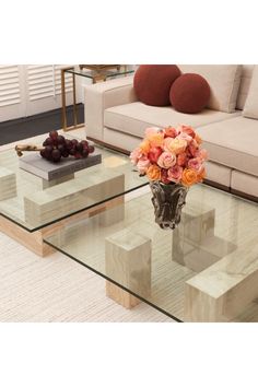 a glass coffee table with flowers and fruit on it in front of a couches