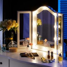 a vanity with lighted mirrors and flowers on it at night, in front of a window