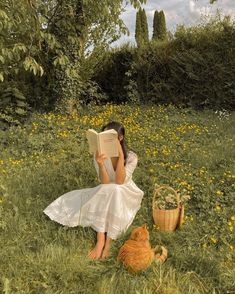 a woman sitting in the grass reading a book with an orange cat laying next to her