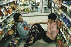 two people sitting on the ground in front of a store filled with food and drinks