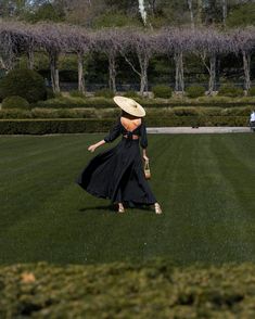 a woman in a long black dress and hat walking across a lush green field