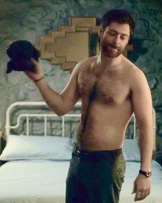 a shirtless man is holding a black object in his right hand while standing next to a bed