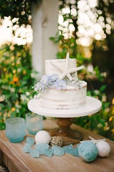 a white wedding cake with blue flowers and seashells on the table next to it