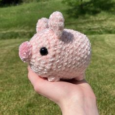 a hand holding a small pink stuffed animal in the middle of a grassy field with trees and grass behind it