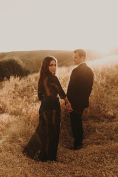 a man and woman holding hands while standing in tall grass with the sun behind them