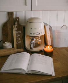 an open book sitting on top of a wooden table next to a coffee maker and candle