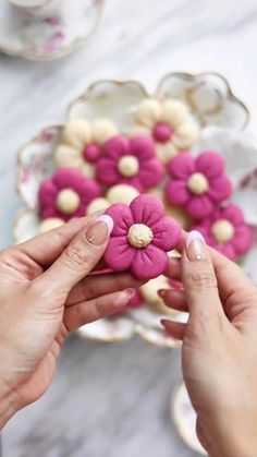 two hands holding pink and white flowers in front of a plate with cookies on it