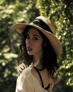 a woman wearing a hat standing in front of some trees with her hand on her shoulder