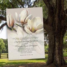 a wedding card with white flowers on it next to a tree in a park area