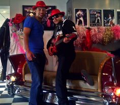 two men standing next to each other in front of a car with pink decorations on it