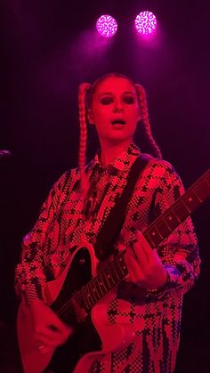 a woman with braids playing an electric guitar on stage at a music festival in front of purple and red lights