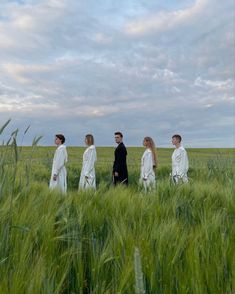 four people dressed in white walking through tall grass on a cloudy day with clouds overhead