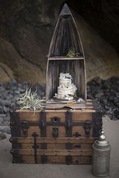 a wedding cake in a wooden box on the beach next to an old trunk with succulents