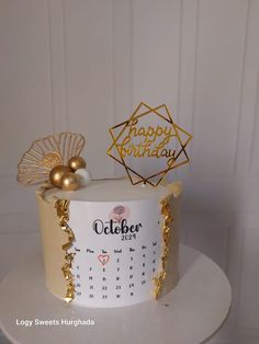 a white and gold birthday cake with a happy birthday calendar on the top, sitting on a table